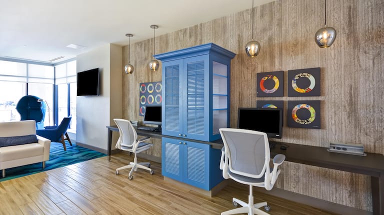 Wall Art, Blue Storage Cabinet Between Two Computers on Long Desk, Two White Ergonomic Chairs in Business Center With TV, Soft Seating, and Large Window in Background