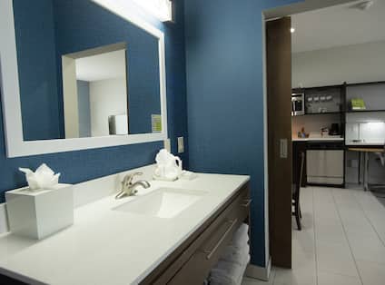 Brightly Lit Vanity Mirror, Sink, Fresh Towels, Toiletries, Amenities in Bathroom With Open Doorway to View of Kitchen and Work Desk