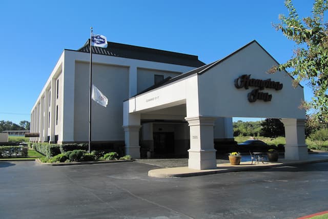 Daytime View of Hotel Exterior With Signage on Porte Cochère, Flagpoles, and Landscaping