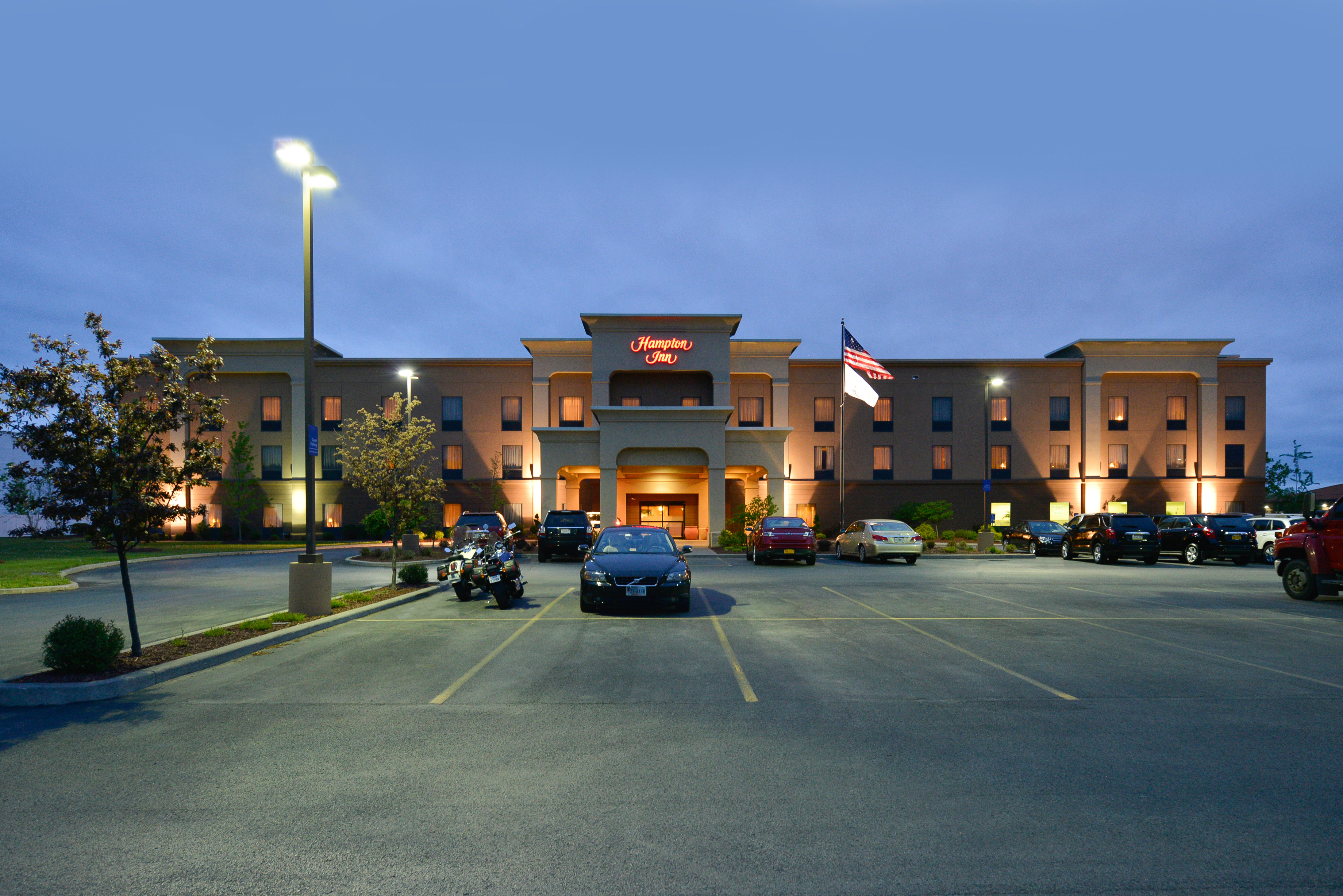 Illuminated Hotel Exterior, Signage, Porte Cochère, Flagpoles, Landscaping, and Guest Cars on Parking Lot at Night