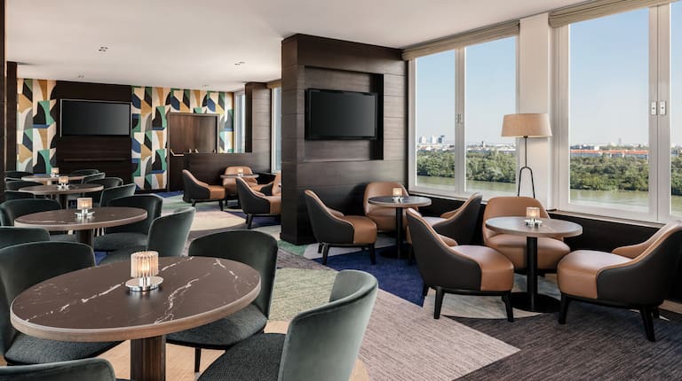 Executive lounge with small round tables and barrel-type chairs along with multiple mounted screens and a wall full of windows.