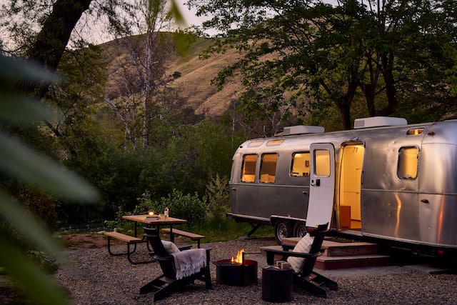 Airstream on right at dusk with outdoor seating and lit fire pit