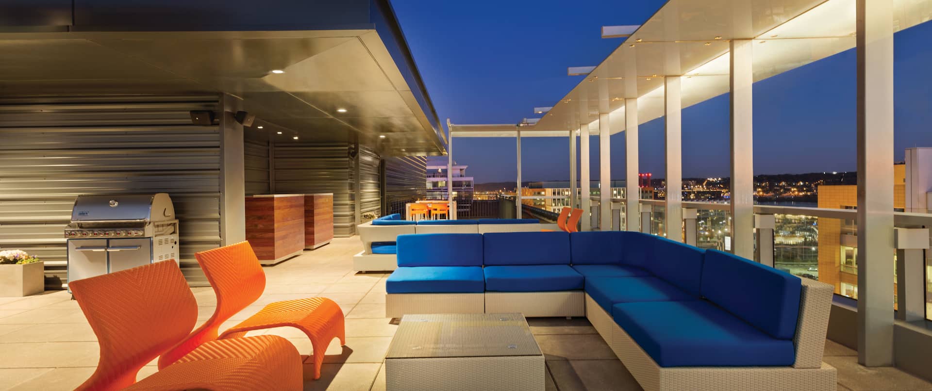 Illuminated Roof Top Terrace With Orange Chairs, Tables, Blue Sofa, and Grill at Night