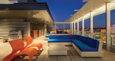 Illuminated Roof Top Terrace With Orange Chairs, Tables, Blue Sofa, and Grill at Night
