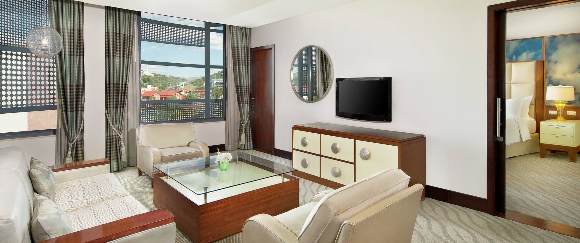Sofa, Stuhl, Tisch und Fernseher in der Diplomatensuite mit offenem Durchgang und Blick auf das Kingsize-Bett