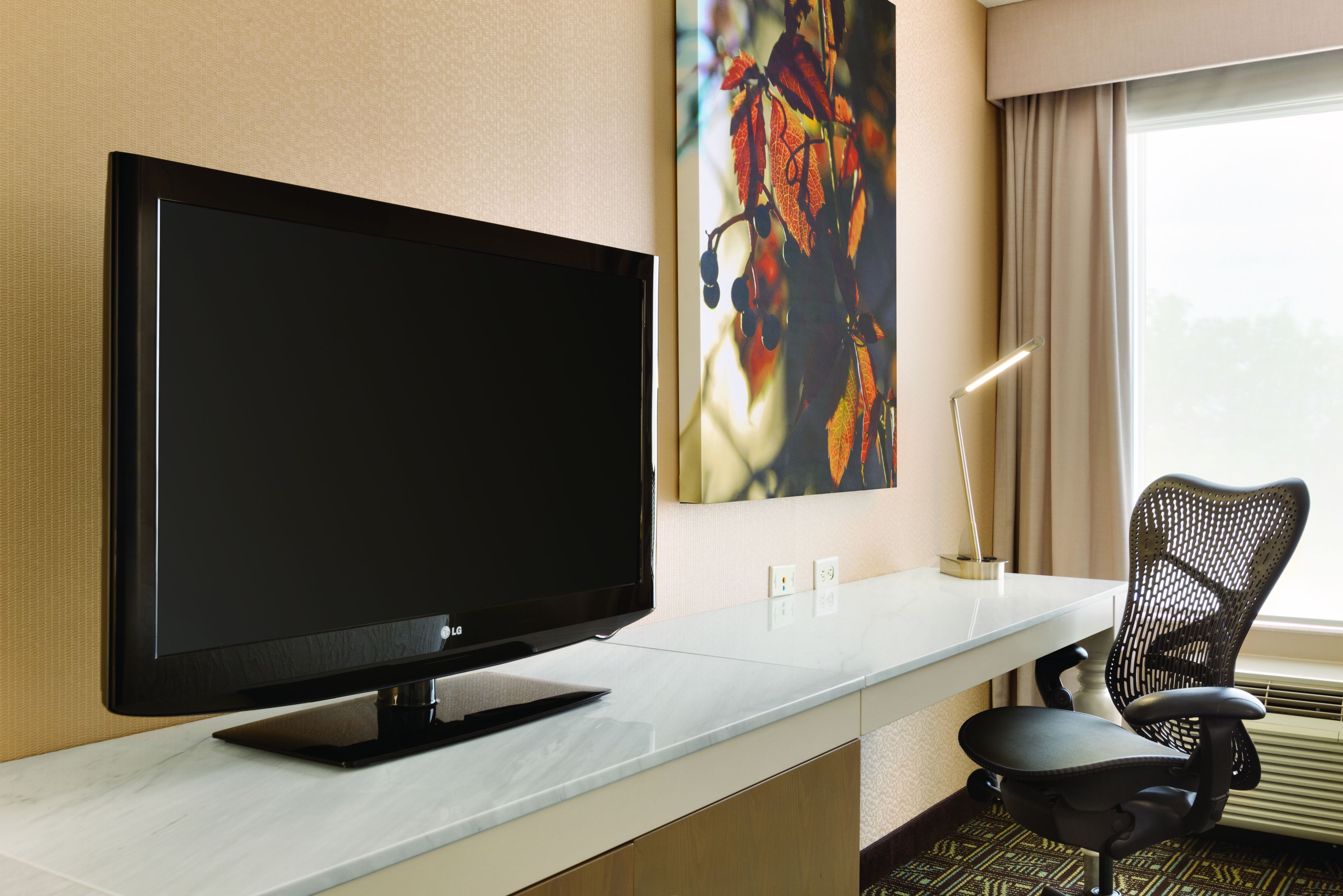 TV, Wall Art, and Desk With Ergonomic Chair by Window With Open Drapes