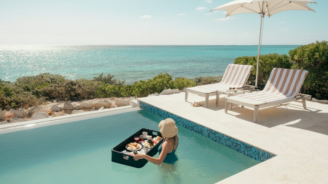 Frau im Swimmingpool am Meer hält schwebendes Frühstück auf dem Tablett