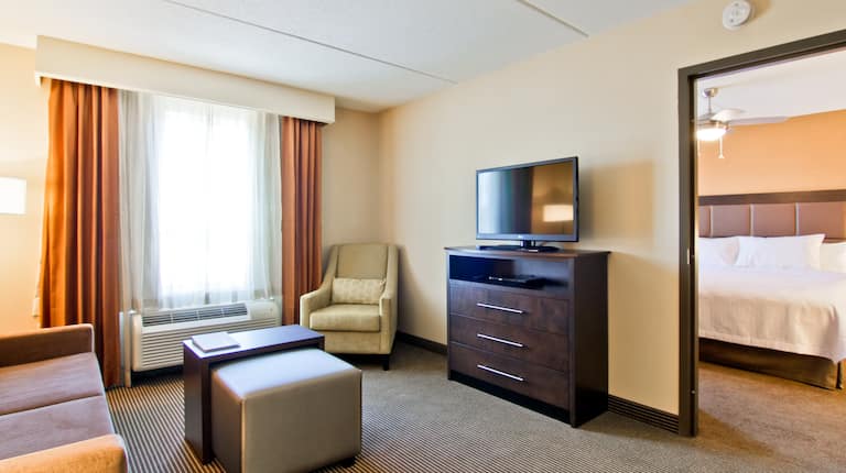 Salle de séjour d'une suite avec canapé, combiné table/pouf, fauteuil, téléviseur et porte ouverte sur la chambre