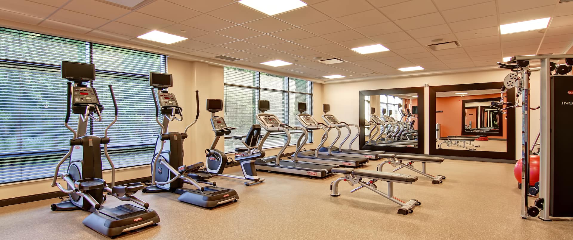 Fitness Room With Cardio Equipment Facing Large Windows, Weight Benches, Two Large Mirrors, Red Exercise Ball, and Weight Machine