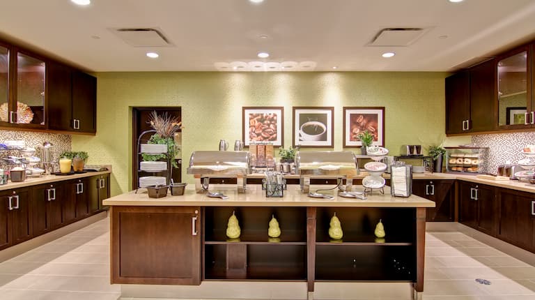 Assiettes, ustensiles, aliment et condiments sur des comptoirs dans le coin repas de l'hôtel avec placards en bois et œuvre d'art au mur