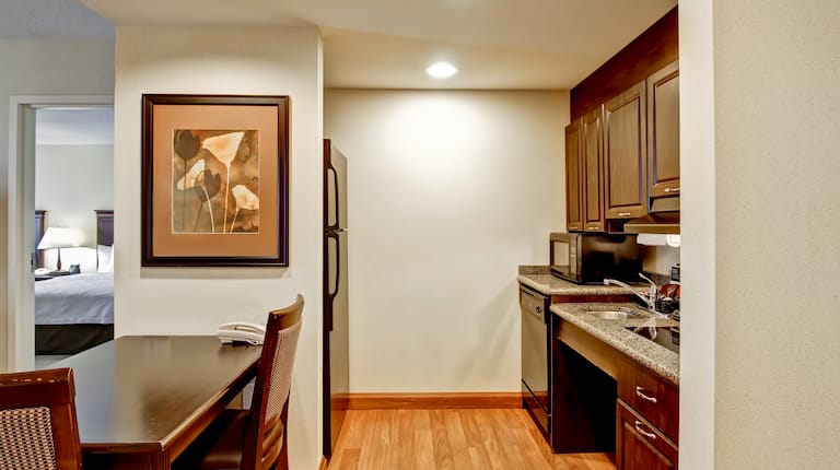 Accessible Kitchen With Fridge, Microwave, Dishwasher, Sink, Wall Art Above Dining Table and Open Doorway to Bedroom