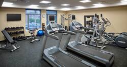 Fitness Center With Weight Bench, TV Above Free Weights, Blue Exercise Ball, Weight Balls, Wall Clock, and Cardio Equipment Facing Windows With Outside View 